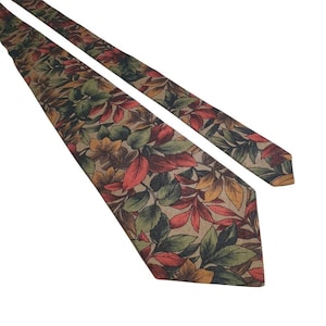 May include: A necktie featuring a repeating pattern of autumn-colored leaves in shades of red, orange, yellow, and dark green set against a tan background. The tie has a pointed tip and a long, narrow shape with a visible texture throughout the fabric.