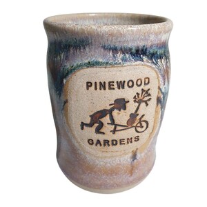 May include: A cylindrical ceramic mug with a speckled beige and blue-toned glaze. The front features a recessed rectangular panel with the text PINEWOOD GARDENS and a stamped illustration of a person pushing a wheelbarrow with a small tree.