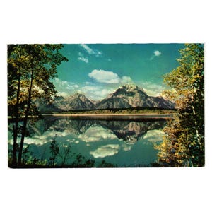 May include: Vintage postcard featuring a scenic mountain landscape reflected in a calm lake. The image showcases snow-capped peaks, fluffy white clouds, and trees with green and yellow foliage under a blue sky. The postcard has a slightly aged appearance.