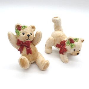 May include: Two ceramic teddy bear figurines, one sitting with its arms raised and the other standing on all fours. Both bears are decorated with a red bow and a green holly wreath.
