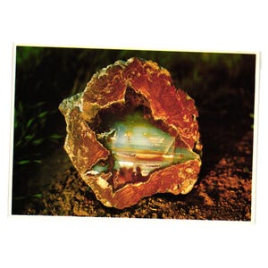 May include: A close-up of a polished, raw geode with a rough, reddish-brown exterior. The interior reveals layers of translucent, light blue, and green crystal formations. The geode sits on a dark brown surface.