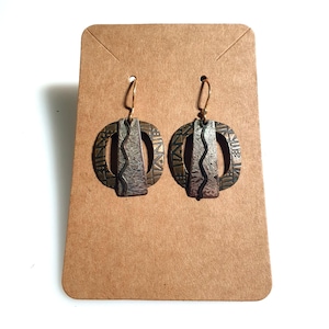 May include: A pair of bronze-toned earrings with a central silver-colored vertical element. The earrings have a circular design with etched patterns and gold-colored ear wires. The earrings are displayed on a brown card.