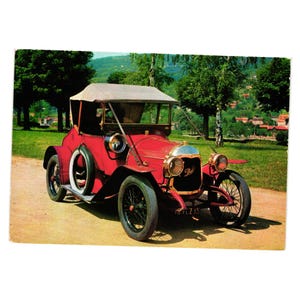 May include: A vintage red car with black wheels and a tan convertible top is parked on a dirt road. The car has a chrome front and headlights. The background features green trees and a glimpse of a village.