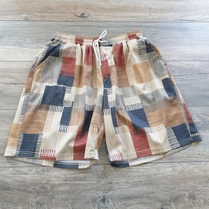 May include: A pair of shorts featuring a patchwork pattern of rectangular blocks in shades of tan, brown, dark blue, and cream. The shorts have an elastic waistband with a white drawstring cord and a textured fabric finish throughout the garment.