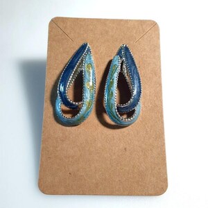 May include: A pair of blue and teal teardrop earrings with silver-colored trim. The earrings have a layered design with a marbled effect and are displayed on a brown card.