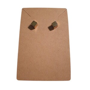 May include: A pair of small, gold-colored cube-shaped stud earrings attached to a rectangular piece of light brown cardboard display paper. The earrings feature a smooth, metallic finish and are positioned side by side on the display card.