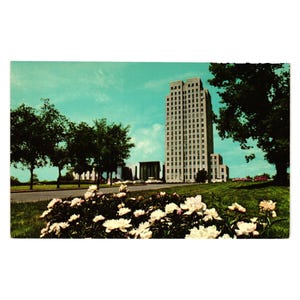 May include: Vintage postcard featuring a tall, multi-story building with numerous windows, set against a backdrop of a blue sky with clouds. The foreground showcases a vibrant display of white flowers, with trees lining the road.