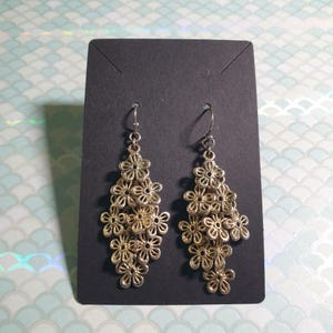 Puede incluir: Un par de pendientes colgantes dorados. Cada pendiente presenta una cascada de pequeñas formas de flores caladas. Los pendientes se exhiben en una tarjeta negra sobre un fondo con un patrón azul claro y blanco.