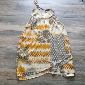 May include: A halter-neck top with a patchwork design. The top features a mix of patterns in shades of yellow, white, and brown. The fabric appears to be lightweight, with a flowing silhouette. The top has a keyhole neckline and a tie closure at the neck.