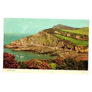 May include: Vintage postcard featuring a coastal landscape with a rocky cliff, green hills, and a turquoise bay. The postcard shows a view of Combe Martin, with boats in the water and lush vegetation in the foreground. The sky is a light blue.