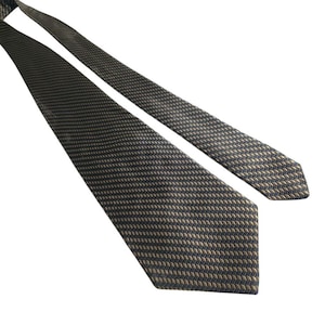 Pode incluir: Uma gravata cinza escuro com um padrão repetitivo de pequenas formas marrons claras. A gravata está em ângulo sobre um fundo branco, mostrando sua superfície texturizada e seu design. A extremidade pontiaguda da gravata é visível.
