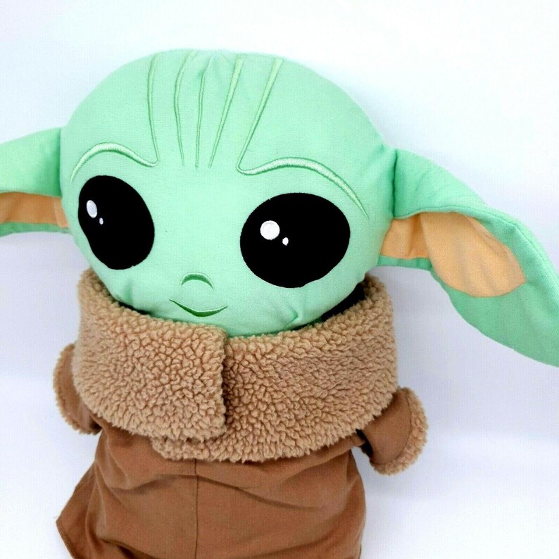 Puede incluir: Mu&ntilde;eco de peluche de un personaje con grandes ojos negros, una cara verde claro y orejas grandes. El mu&ntilde;eco lleva un abrigo marr&oacute;n con un cuello difuso de color canela. La cara del mu&ntilde;eco tiene una peque&ntilde;a sonrisa curvada.