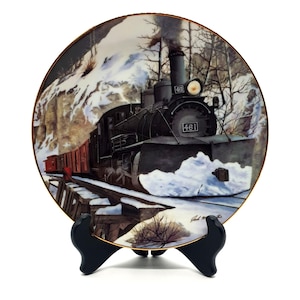 May include: A decorative plate featuring a black steam train with the number 461, traveling through a snowy landscape. The plate has a gold rim and is displayed on a black stand. The train is pulling red train cars.