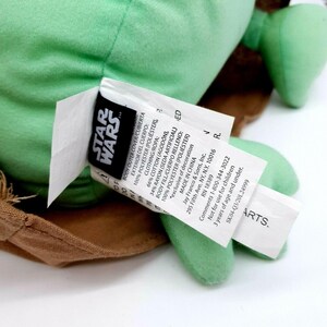 Puede incluir: Un peluche verde con una t&uacute;nica marr&oacute;n y una etiqueta que dice "STAR WARS". La etiqueta incluye detalles sobre los materiales, incluyendo poli&eacute;ster y algod&oacute;n. El juguete no es para ni&ntilde;os menores de 3 a&ntilde;os.
