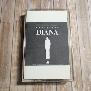 Può includere: Una musicassetta vintage con l'album "Anthology Diana". La copertina è in bianco e nero, con il nome "DIANA" a grandi lettere bianche e la silhouette di una persona. La cassetta è in un astuccio di plastica trasparente.