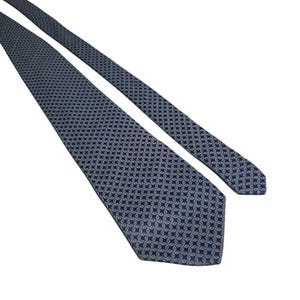 Puede incluir: Una corbata azul oscuro con un patrón geométrico repetitivo de pequeñas formas ovaladas entrelazadas en azul claro y dorado. La corbata tiene un extremo puntiagudo y un cuerpo largo y estrecho, mostrando un diseño intrincado y uniforme.