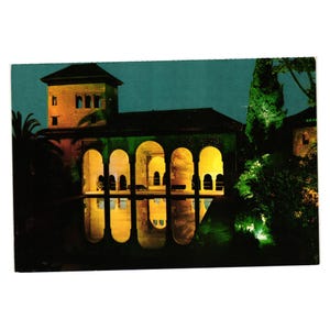 May include: A vintage postcard featuring a nighttime view of a building with arched windows reflected in water. The building is illuminated with warm yellow light, contrasting with the dark blue-green sky. Palm trees and other foliage frame the scene.
