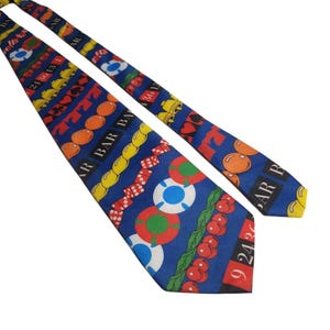 May include: A blue necktie featuring a repeating casino-themed pattern. The design includes illustrations of red dice, poker chips in red, green, and blue, gold bars, cherries, bells, playing card suits, and roulette wheel numbers in black and red.