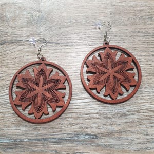 Peut inclure: Une paire de boucles d'oreilles en bois brun avec un motif de fleur sculpté. Les boucles d'oreilles sont rondes et ont un crochet argenté.