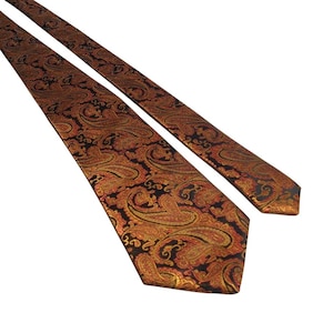 May include: A patterned tie with a paisley design in shades of gold, orange, and black. The tie is made of a shiny material and is angled diagonally across the frame. The paisley pattern is detailed and covers the entire surface of the tie.