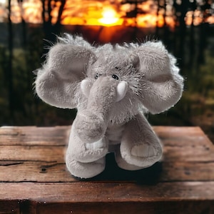 May include: A plush gray elephant toy with large ears, a trunk, and white tusks. The stuffed animal is sitting on a wooden surface. The background features a blurred view of trees and a sunset.