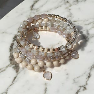 May include: A set of five layered bracelets featuring a mix of cream, white, and light pink beads with gold accents. The bracelets have two heart-shaped charms.