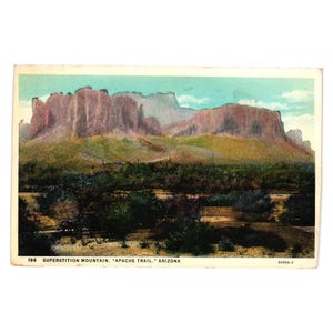 May include: Vintage postcard featuring a landscape of Superstition Mountain in Arizona. The image shows a mountain range with reddish-brown rock formations, green vegetation, and a blue sky. The text on the postcard reads "Superstition Mountain, Apache Trail, Arizona."