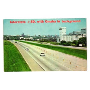 May include: A vintage postcard showing a multi-lane highway with cars driving on Interstate 80. The text Interstate # 80, with Omaha in background is printed at the top. The scene features green grassy medians and a large industrial grain elevator.