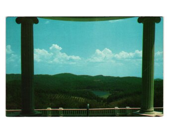 May include: Vintage postcard featuring a scenic landscape viewed through a classical architectural frame. The image showcases a vibrant blue sky with fluffy clouds, overlooking lush green hills and a body of water.