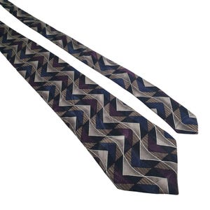 May include: A patterned necktie with a repeating geometric design. The tie features a chevron pattern in shades of navy blue, purple, gray, and tan. The tie is angled across a white background.