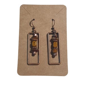 May include: A pair of copper-colored rectangular earrings with a dangle design. Each earring features a series of brown and yellow beads within the frame. The earrings are displayed on a brown card.