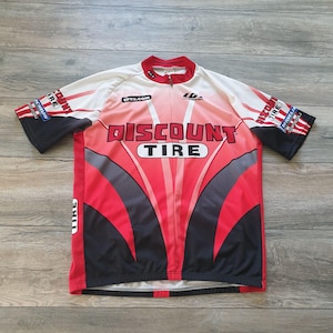 May include: A red, black, and white cycling jersey with the words "Discount Tire" prominently displayed on the front. The jersey has a full zipper, short sleeves, and the tires.com logo. The jersey is made of a lightweight, breathable material.