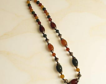 Puede incluir: Un collar de cuentas con tonos tierra. Presenta cuentas ovaladas y redondas en tonos marrón, naranja, verde y amarillo, intercaladas con elementos metálicos plateados. El collar se exhibe sobre una superficie clara, resaltando sus colores.