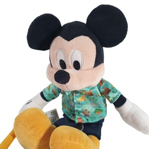 May include: A plush Mickey Mouse toy wearing a green and blue Hawaiian shirt with a pattern of Disney theme park attractions. The toy has black ears, a black nose, and yellow hands and feet.