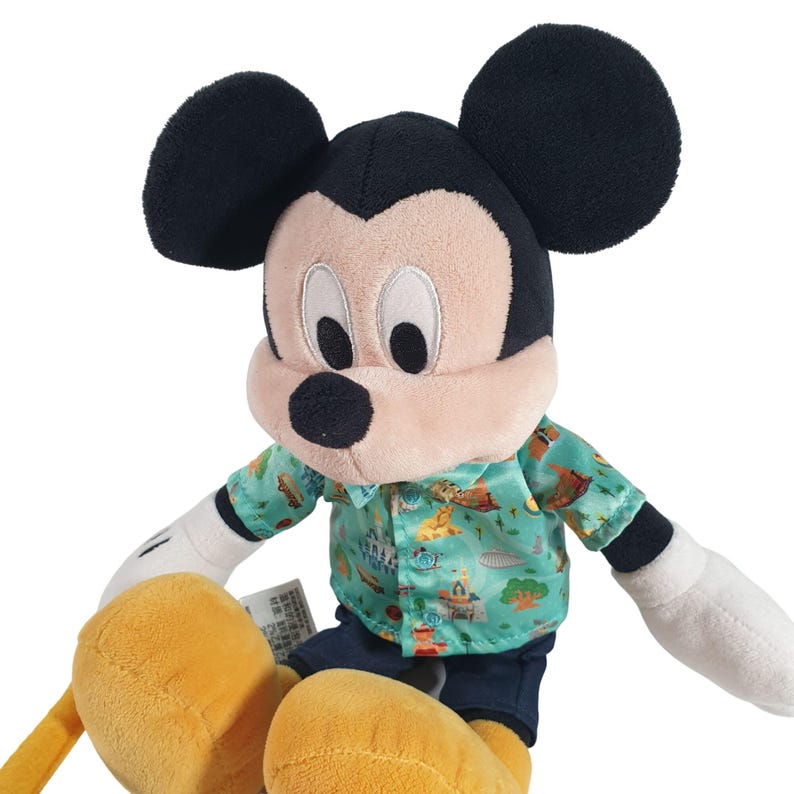 May include: A plush Mickey Mouse toy wearing a green and blue Hawaiian shirt with a pattern of Disney theme park attractions. The toy has black ears, a black nose, and yellow hands and feet.