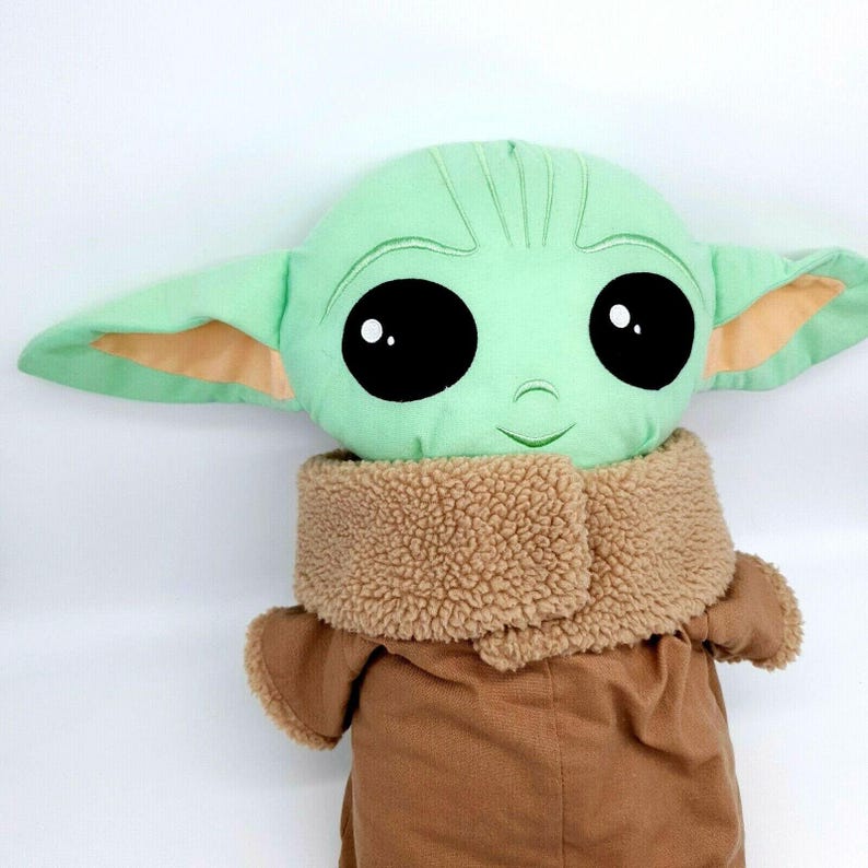 Puede incluir: Un mu&ntilde;eco de peluche de un personaje con grandes ojos negros, piel verde y orejas grandes. El mu&ntilde;eco lleva un abrigo marr&oacute;n con un cuello lanudo color canela. La cabeza del mu&ntilde;eco es verde claro con detalles bordados.