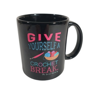 May include: Black ceramic mug with a white and pink design that says "Give yourself a crochet break." The mug has a crochet hook and yarn balls in a basket.