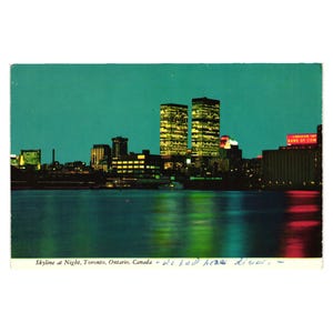 May include: A vintage postcard featuring the Toronto, Ontario, Canada skyline at night. The image displays illuminated skyscrapers reflecting on dark water, with a red sign reading CANADIAN IMPERIAL BANK OF COMMERCE and handwritten text.