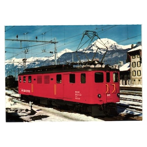 May include: A vintage postcard featuring a bright red electric train against a backdrop of snow-capped mountains and a clear blue sky. The train has the number 911 and the text "SBB CRIFFS" on its side. Power lines and buildings are also visible.