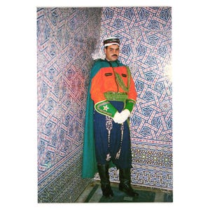 Puede incluir: Una persona con un uniforme militar marroquí tradicional. El uniforme incluye una chaqueta naranja, una capa verde, pantalones azules y botas negras. El fondo presenta una pared de azulejos estampados.