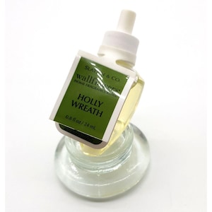 May include: A clear glass air freshener bottle with a white top and a green and white label that reads "HOLLY WREATH". The bottle contains a yellow liquid and is 0.8 fl oz / 24 mL.