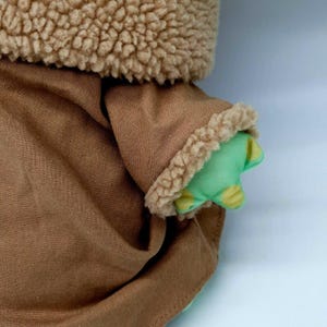 Puede incluir: Primer plano de la ropa de tela marr&oacute;n de un juguete de peluche con un ribete difuso marr&oacute;n. La mano verde del juguete es visible, con manchas amarillas. La ropa del juguete est&aacute; hecha de un material suave y texturizado.