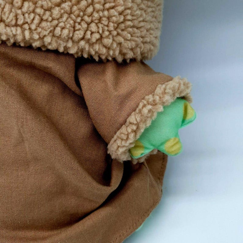 Puede incluir: Primer plano de la ropa de tela marr&oacute;n de un juguete de peluche con un ribete difuso marr&oacute;n. La mano verde del juguete es visible, con manchas amarillas. La ropa del juguete est&aacute; hecha de un material suave y texturizado.