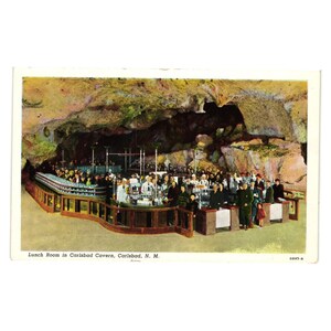 May include: Vintage postcard featuring the Lunch Room in Carlsbad Cavern, New Mexico. The image shows a large group of people inside a cave, gathered around a counter. The cave walls are visible in the background, with the text "Lunch Room in Carlsbad Cavern, Carlsbad, N. M."