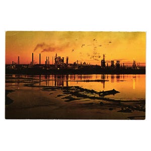 May include: Vintage postcard featuring an industrial landscape at sunset. Silhouetted factories and smokestacks are reflected in the water, under a vibrant orange and yellow sky. The scene evokes a sense of nostalgia.