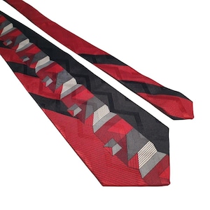 Puede incluir: Una corbata estampada en rojo, negro y gris. La corbata presenta un diseño geométrico con formas rojas, negras y grises. La corbata está en ángulo en el marco, con la punta apuntando hacia la esquina inferior izquierda.