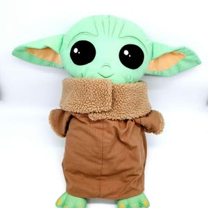 Puede incluir: Un mu&ntilde;eco de peluche de un personaje con grandes ojos negros, piel verde y orejas grandes. El mu&ntilde;eco lleva un abrigo marr&oacute;n con un cuello de piel sint&eacute;tica beige. Los pies y las manos del mu&ntilde;eco son verdes.