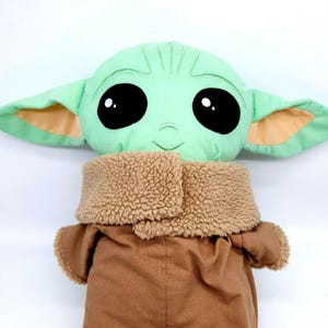 Puede incluir: Un mu&ntilde;eco de peluche de un personaje con grandes ojos negros, piel verde y orejas grandes. El mu&ntilde;eco lleva un abrigo marr&oacute;n con un cuello difuso color canela. El mu&ntilde;eco es un personaje popular de una serie de ciencia ficci&oacute;n.