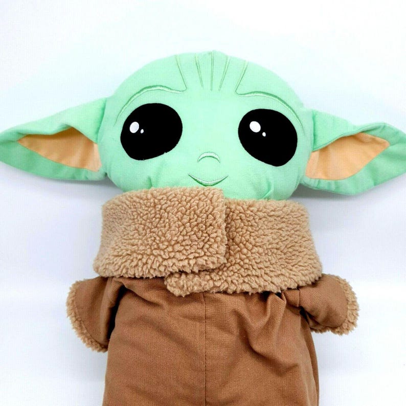 Puede incluir: Un mu&ntilde;eco de peluche de un personaje con grandes ojos negros, piel verde y orejas grandes. El mu&ntilde;eco lleva un abrigo marr&oacute;n con un cuello difuso color canela. El mu&ntilde;eco es un personaje popular de una serie de ciencia ficci&oacute;n.