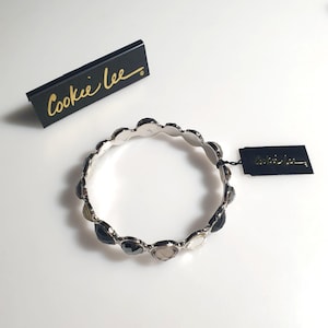 Pode incluir: Uma pulseira prateada com borda ondulada e pedras pretas, cinzentas e transparentes em forma de coração. A pulseira está ao lado de uma etiqueta preta e de uma caixa preta com o texto dourado "Cookie Lee".